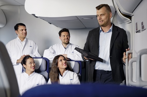 Several people are seated in a cabin mock-up, with a trainer standing beside them