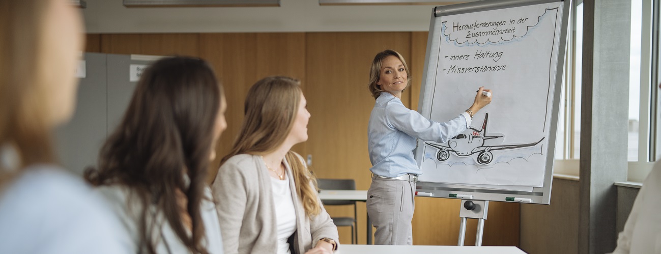 Eine Trainer:in steht an einem Flipchart und notiert etwas, dahinter sitzen Workshopteilnehmer:innen.