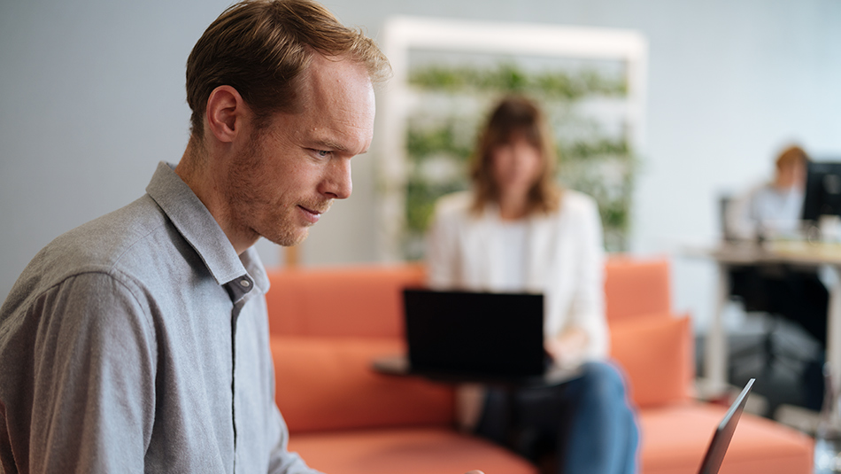 Eine Person im Vordergrund blickt nach unten auf einen Laptop, im Hintergrund eine Person auf einer Couch mit Laptop vor sich.