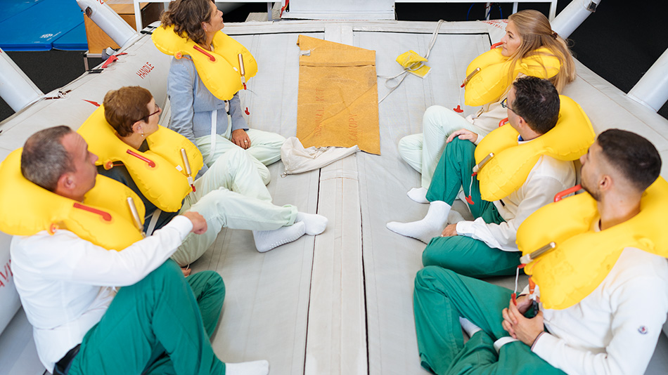 Mehrere Personen sitzen mit Schwimmwesten in einem Rettungsboot auf einem Hallenboden.